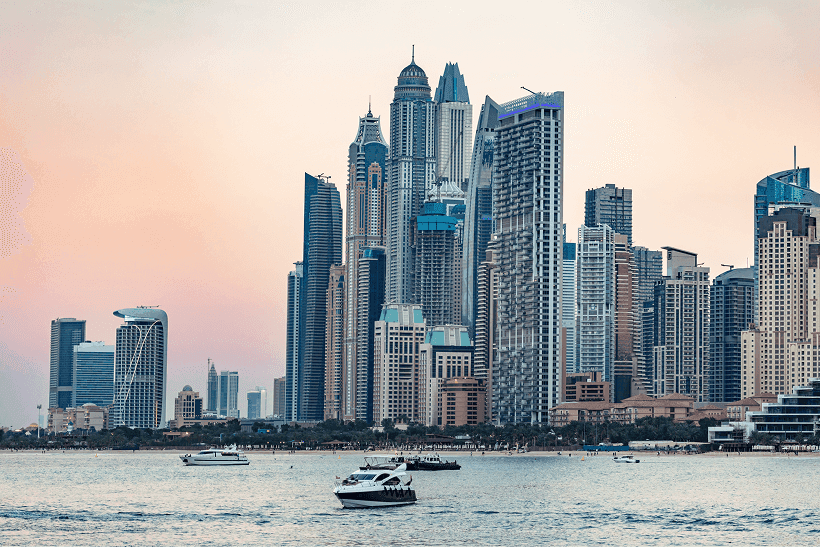 Skyline of modern skyscrapers at sunset, set against a pink and orange sky. Boats are on calm water in the foreground, conveying a serene atmosphere.